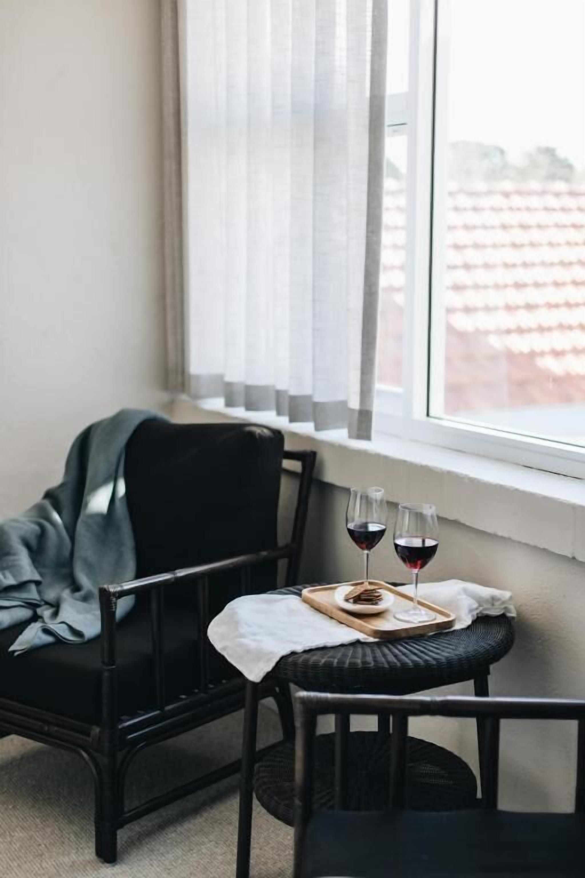 A corner room with a small table set with two glasses of red wine and a tray of snacks, a chair, and a window with sheer curtains.