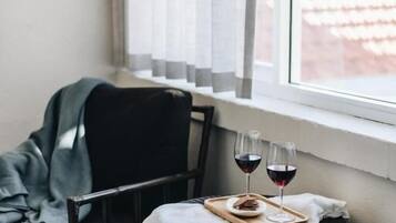 A corner room with a small table set with two glasses of red wine and a tray of snacks, a chair, and a window with sheer curtains.
