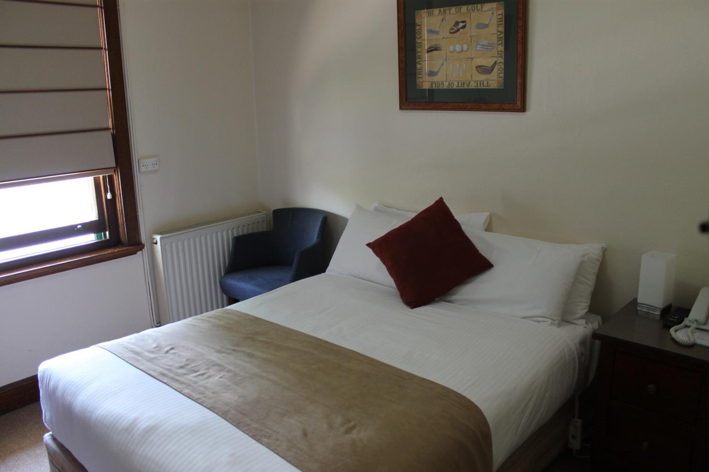 A hotel room with a bed, a chair, a radiator, a framed picture on the wall, and a bedside table with a phone.