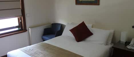 Una habitación de hotel con una cama, una silla, un radiador, un cuadro en la pared y una mesita de noche con un teléfono.