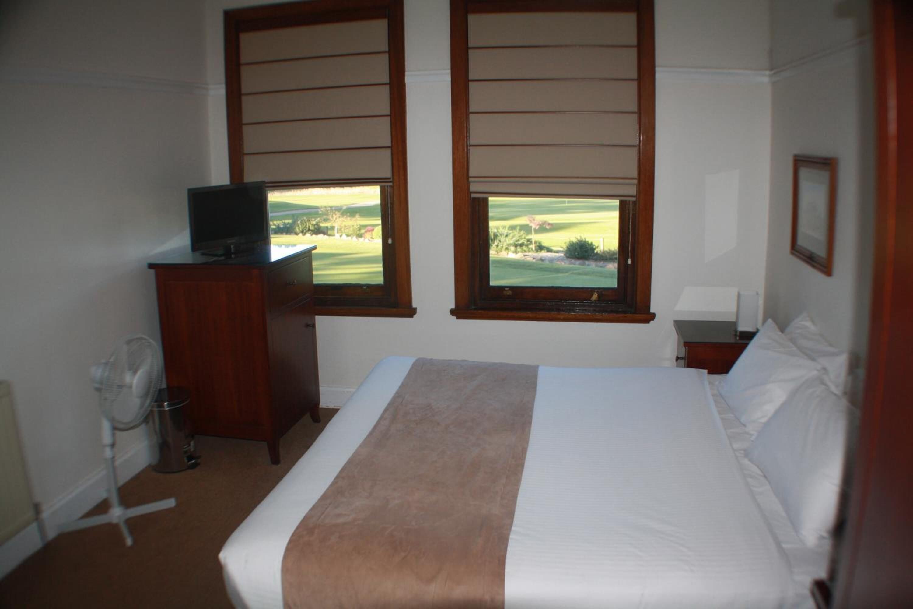 A bedroom with a bed, a television on a wooden cabinet, and two windows with a view of a golf course.