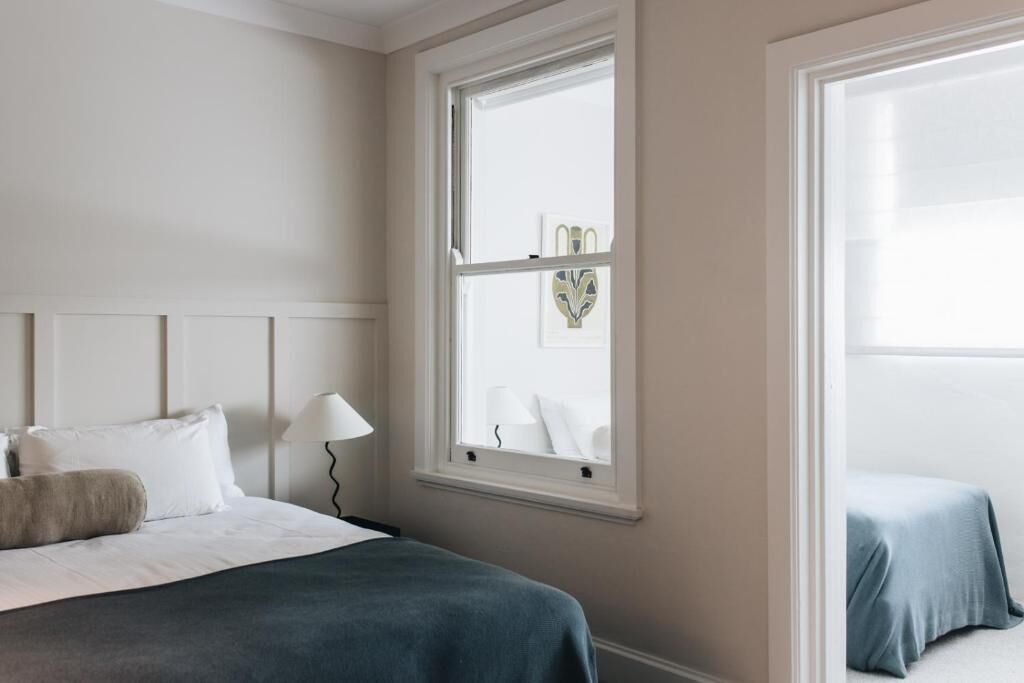 Un dormitorio con cama, mesita de noche, lámpara, ventana y una obra de arte.
