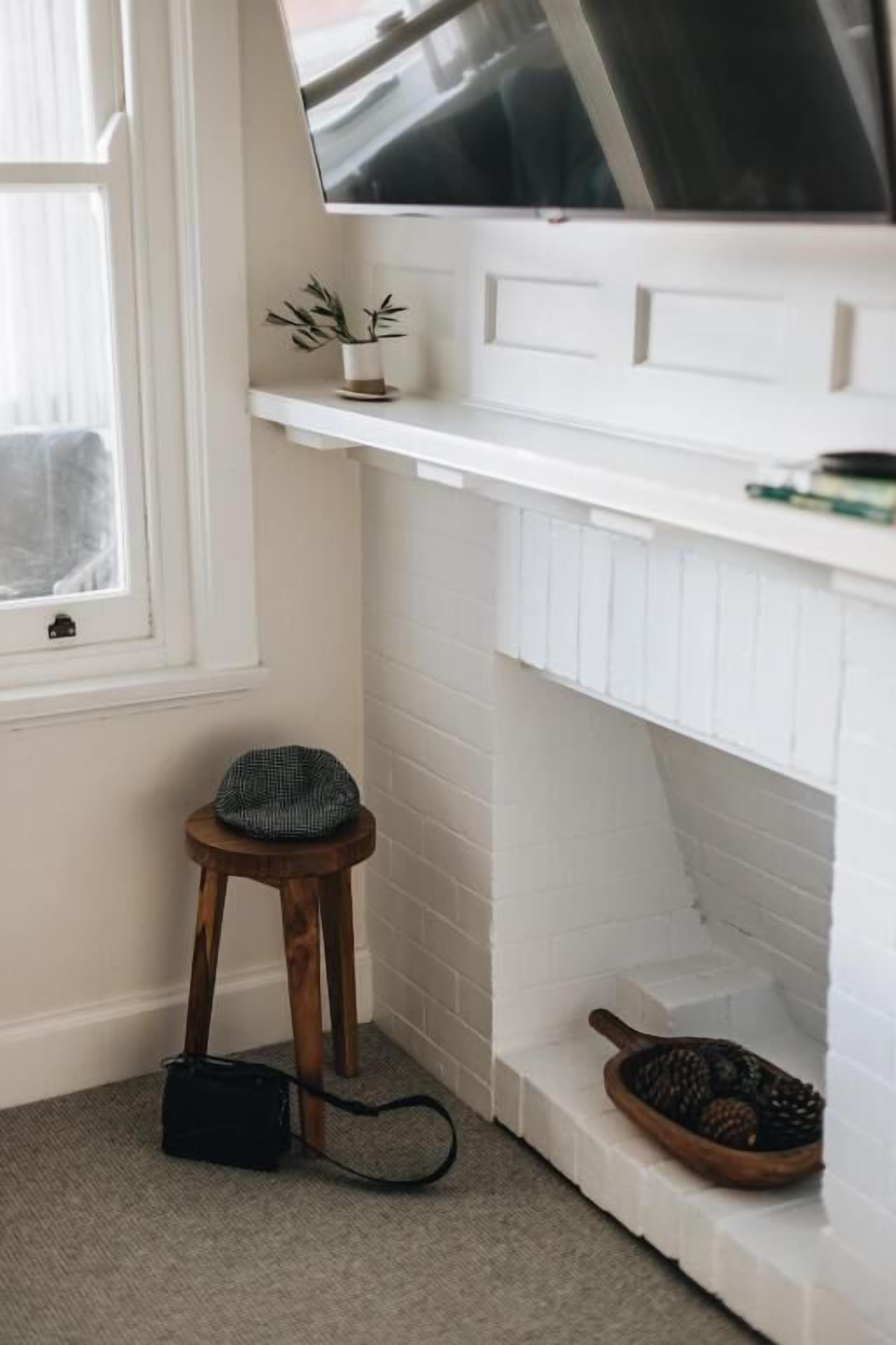 Una esquina de una habitación con una chimenea blanca, un taburete con un sombrero y un bolso negro en el suelo.