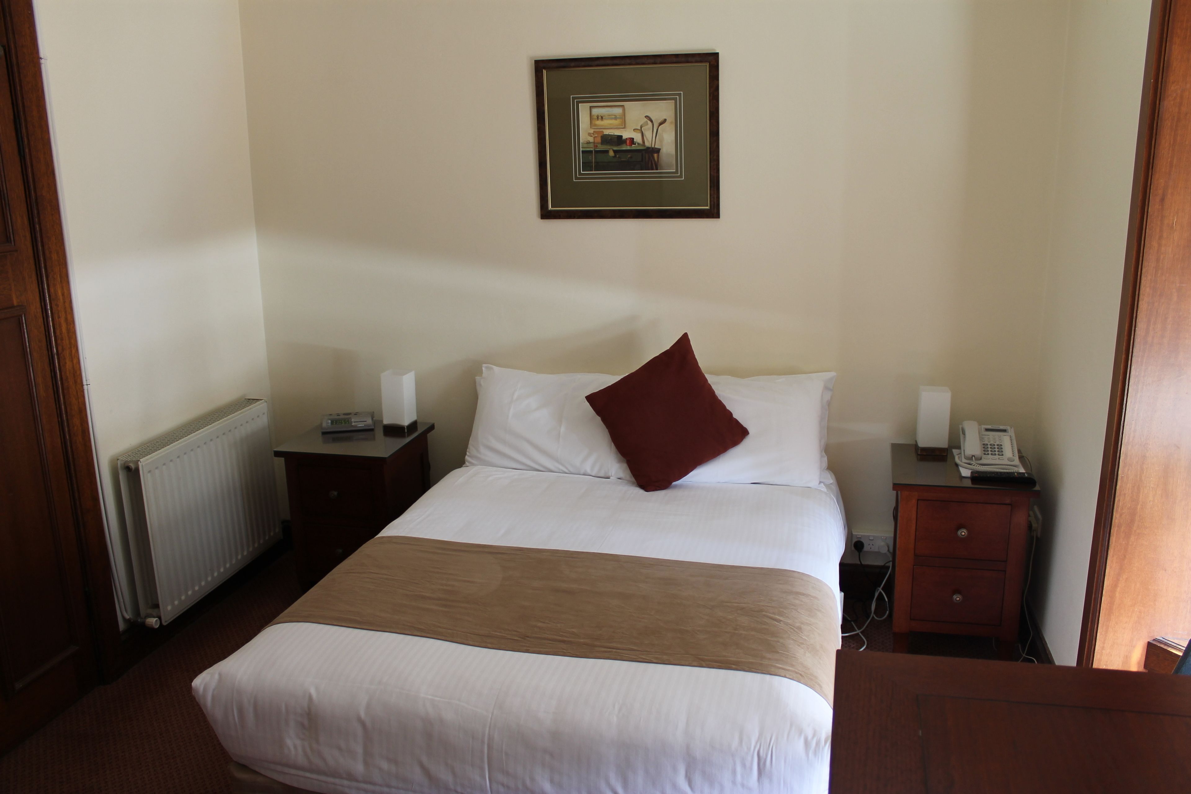 Ein Hotelzimmer mit einem großen Bett, einem Nachttisch mit einer Lampe, einem kleinen Nachttisch mit einem Telefon und einem gerahmten Bild an der Wand.