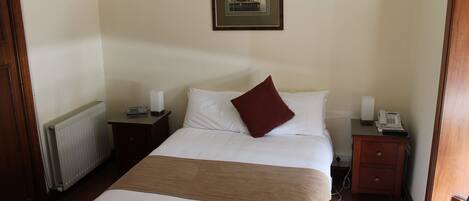 Habitación de hotel con una cama grande, una mesita de noche con lámpara, una mesita más pequeña con teléfono y un cuadro en la pared.