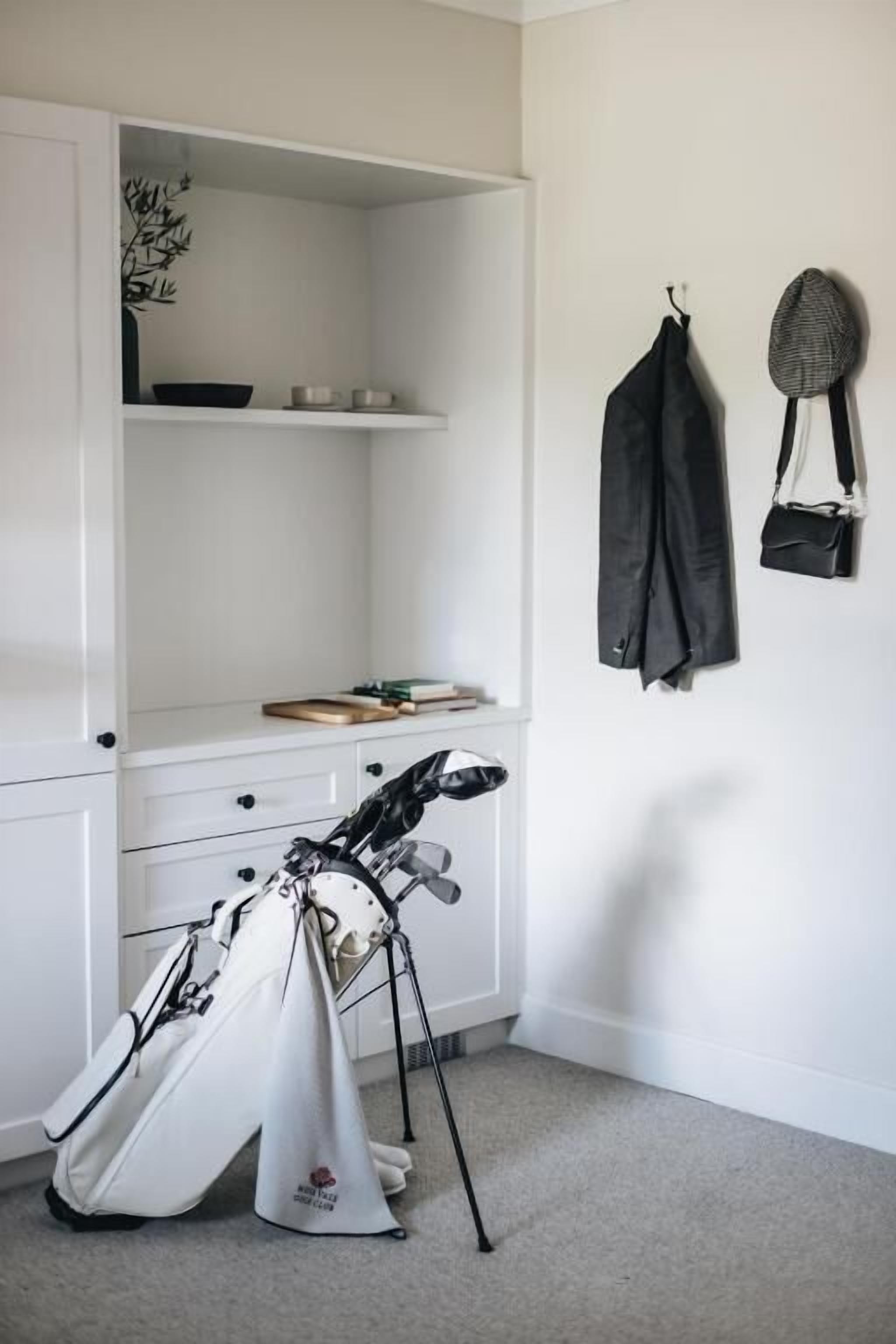 A room with a white wardrobe, a shelf with books and a bowl, a golf bag, and a hat hanging on the wall.