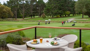 A dining area with wicker furniture, a table set with food and drinks, and a view of a golf course in the background. - The Dormie House - On The Golf Course (Moss Vale)