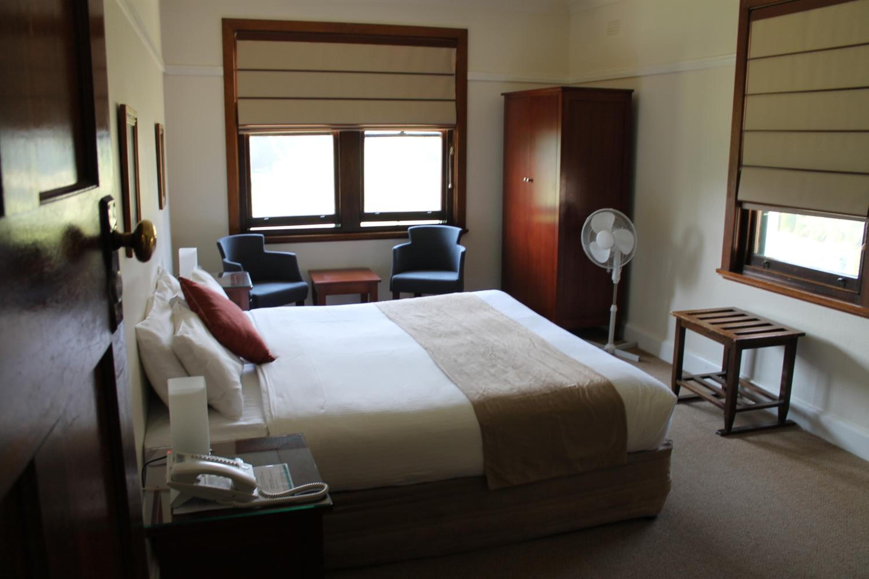 A hotel room with a large bed, two chairs, a desk with a phone, a fan, and a window with blinds.
