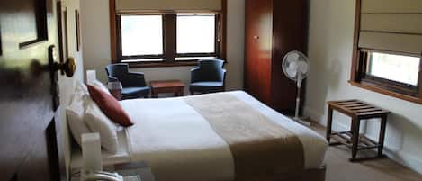 A hotel room with a large bed, two chairs, a desk with a phone, a fan, and a window with blinds.