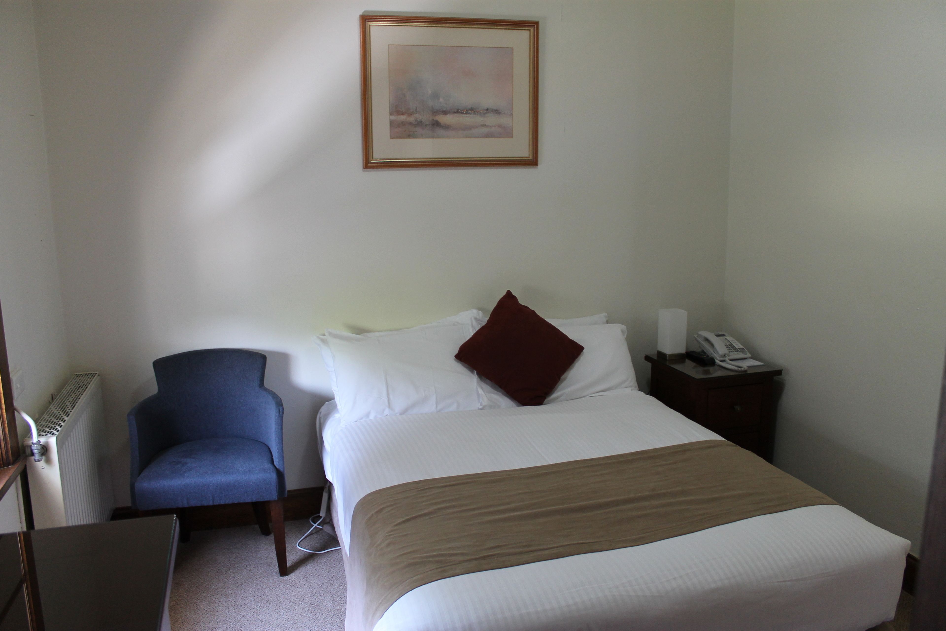 Una habitación de hotel con cama, una silla, mesita de noche con teléfono y un cuadro en la pared.