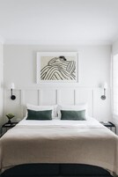 Una cama bien hecha con cabecera blanca y dos almohadas verdes, una obra de arte enmarcada sobre la cama, y una mesita de noche con una lámpara.