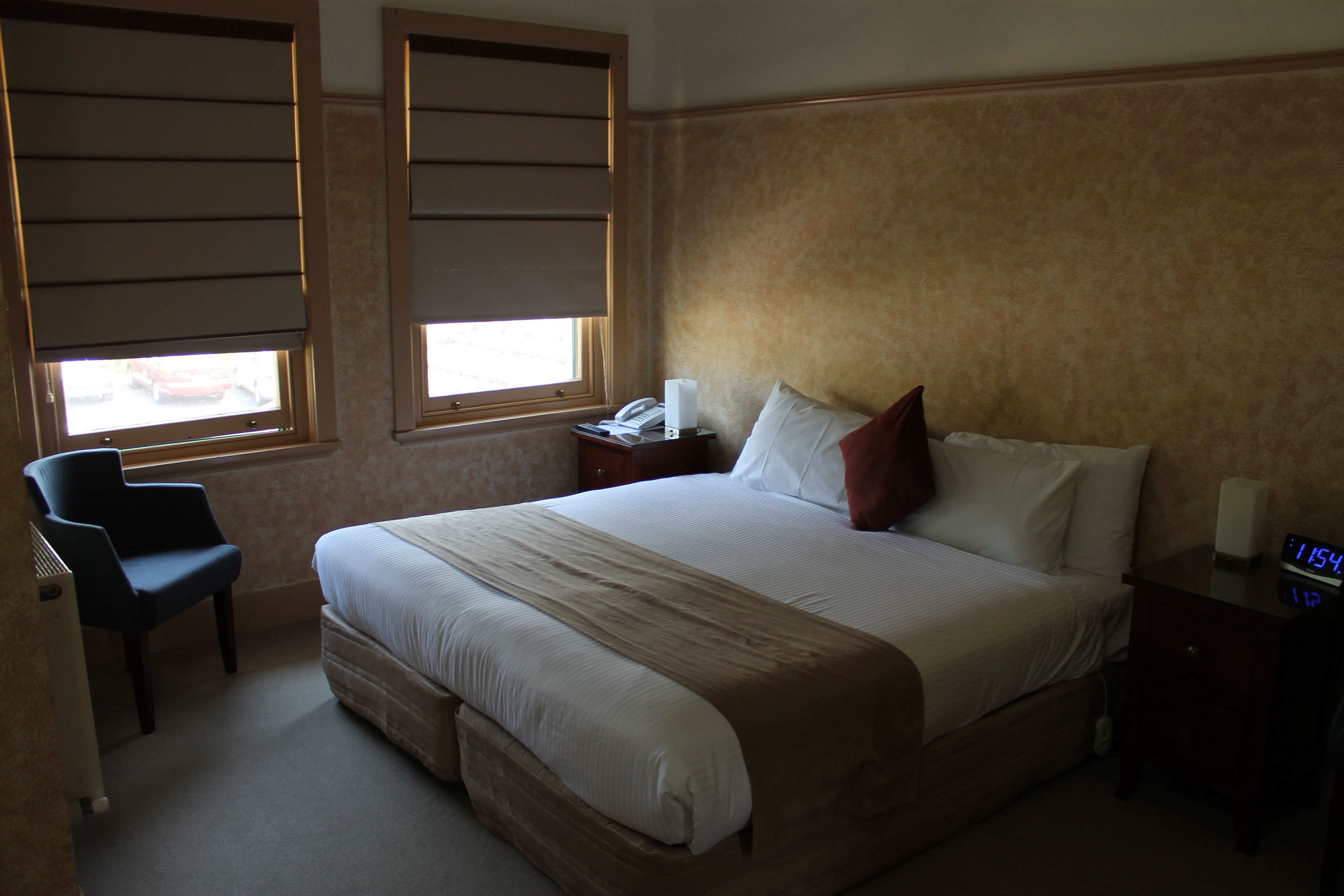 A hotel room with a large bed, a bedside table with a clock, a chair, and a window with blinds.