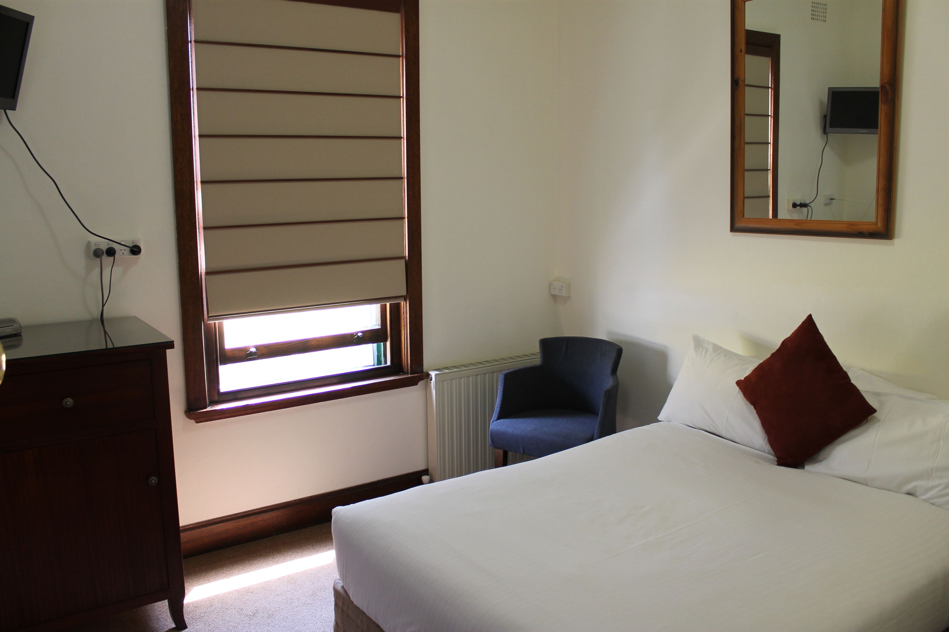 A hotel room with a bed, a chair, a dresser, a window with blinds, and a mirror on the wall.