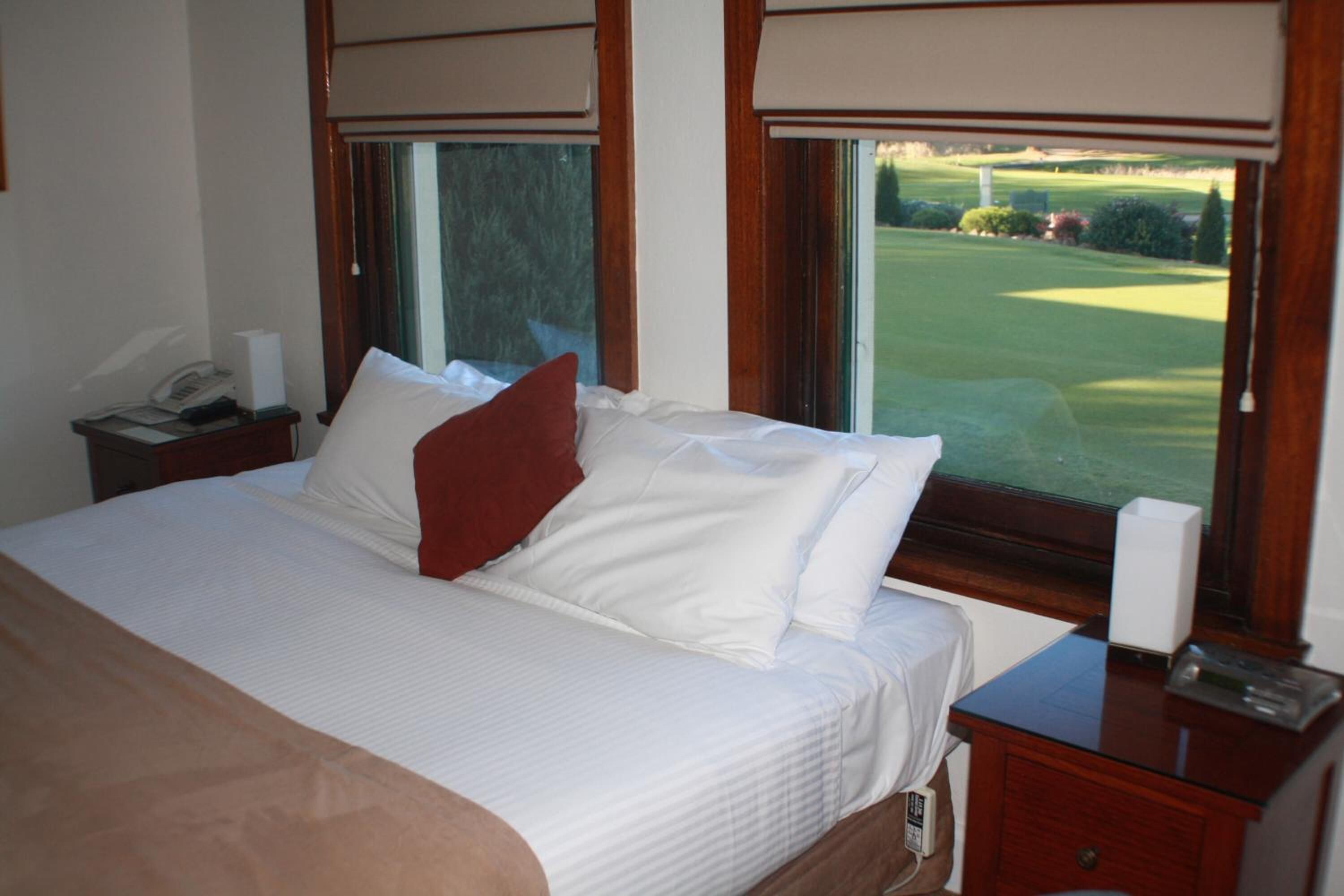 Una cama bien hecha con sábanas blancas y un almohárraga roja, una mesita de noche con un teléfono y una caja de pañuelos, y una ventana con vista a un campo de golf.