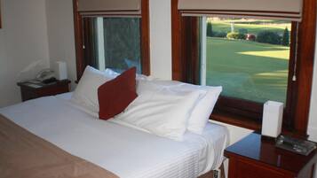 Una cama bien hecha con sábanas blancas y un almohárraga roja, una mesita de noche con un teléfono y una caja de pañuelos, y una ventana con vista a un campo de golf.