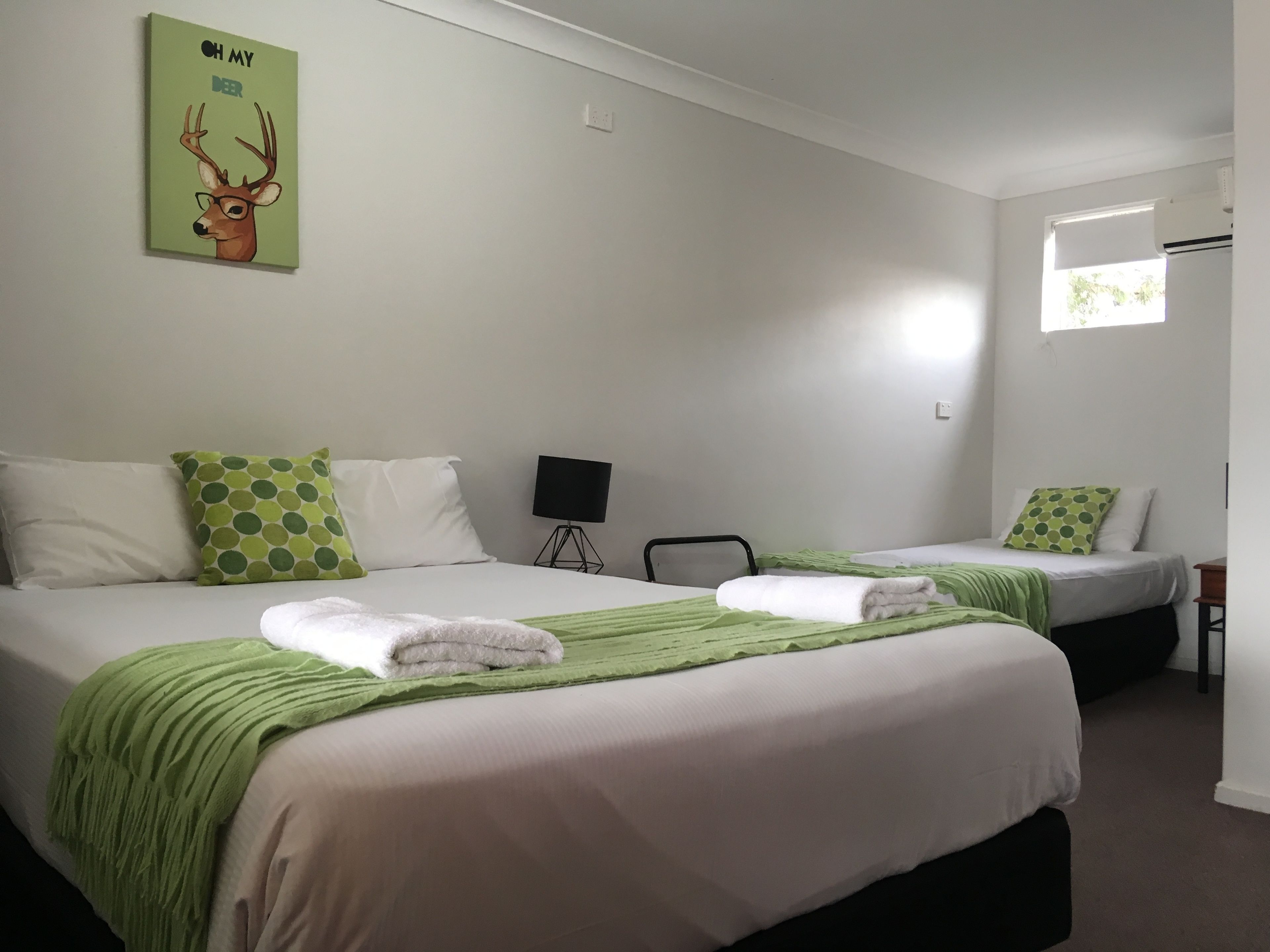 Une chambre d’hôtel avec deux lits, chacun recouvert d’une couverture verte et agrémenté d’oreillers blancs. Un tableau représentant un cerf est accroché au mur, une lampe noire sur trépied se trouve dans un coin, et une fenêtre est dotée de stores.