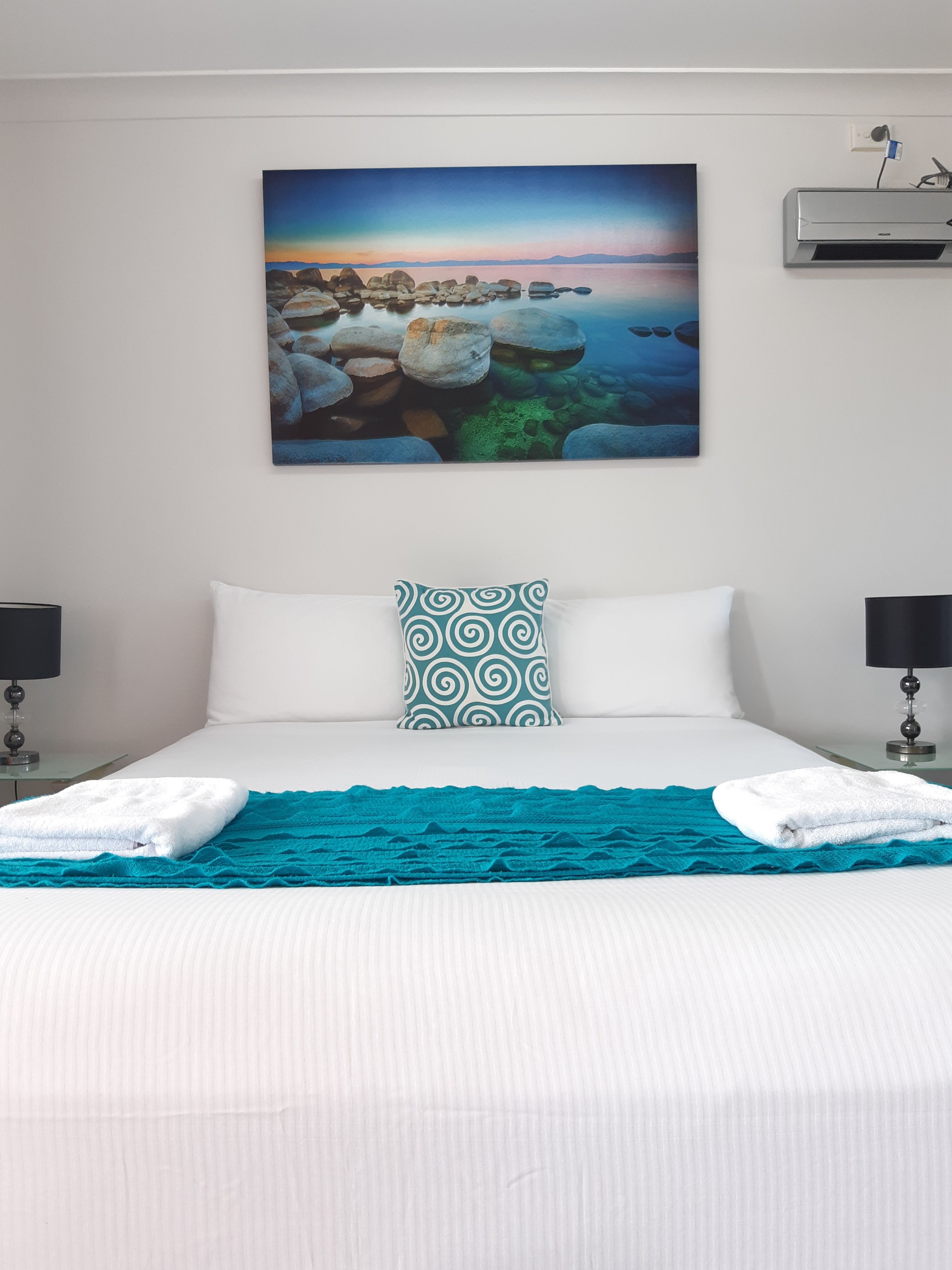 Cama bem arrumada com lençóis brancos, um cobertor azul-turquesa e almofada estampada. Um quadro grande representando uma paisagem costeira está pendurado acima da cama.