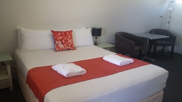 Una cama bien hecha con sábanas blancas, una manta roja y dos toallas dobladas. Hay una almohada con estampado rojo y blanco, una lámpara de noche y una mesita con una silla.