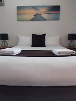 Una cama bien hecha con sábanas blancas y un almohadón negro, flanqueada por dos mesitas de noche con lámparas, en una habitación con vista al atardecer en la pared.