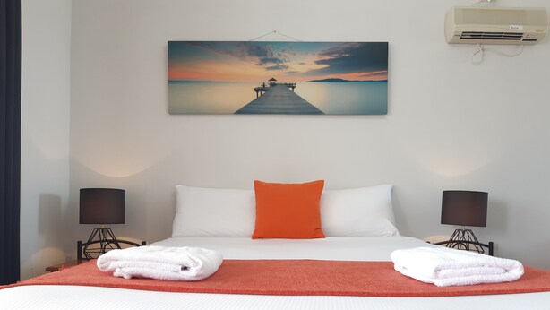 A hotel room with a bed, two bedside lamps, a painting of a sunset over water, and an air conditioning unit. - Econo Lodge Chaparral Motel Ballina (West Ballina)