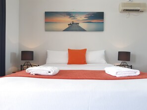 Een hotelkamer met een bed, twee bedlampen, een schilderij van een zonsondergang boven water en een airconditioner.