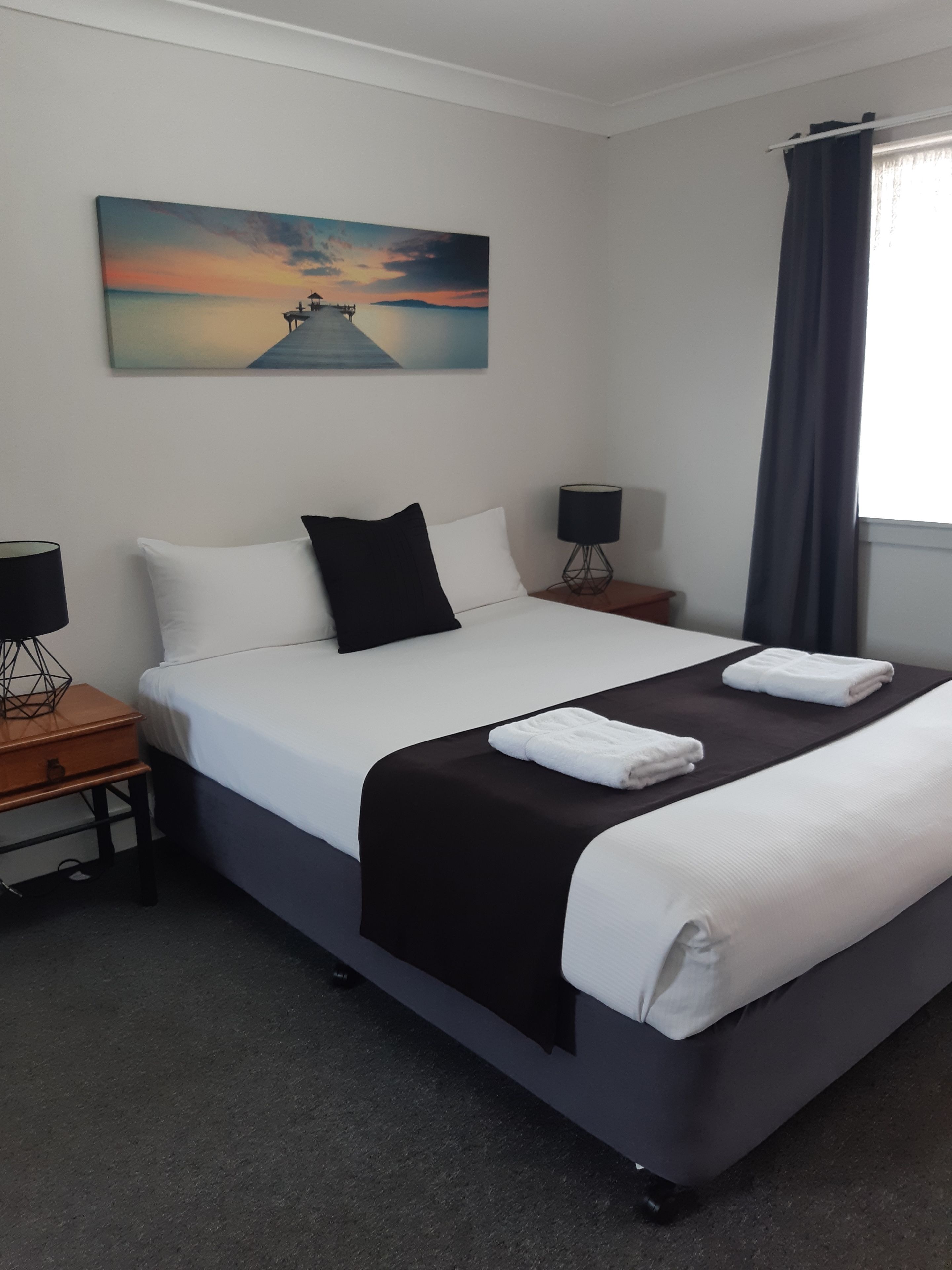 A hotel room with a large bed, two bedside tables, a painting on the wall, and a window with curtains.
