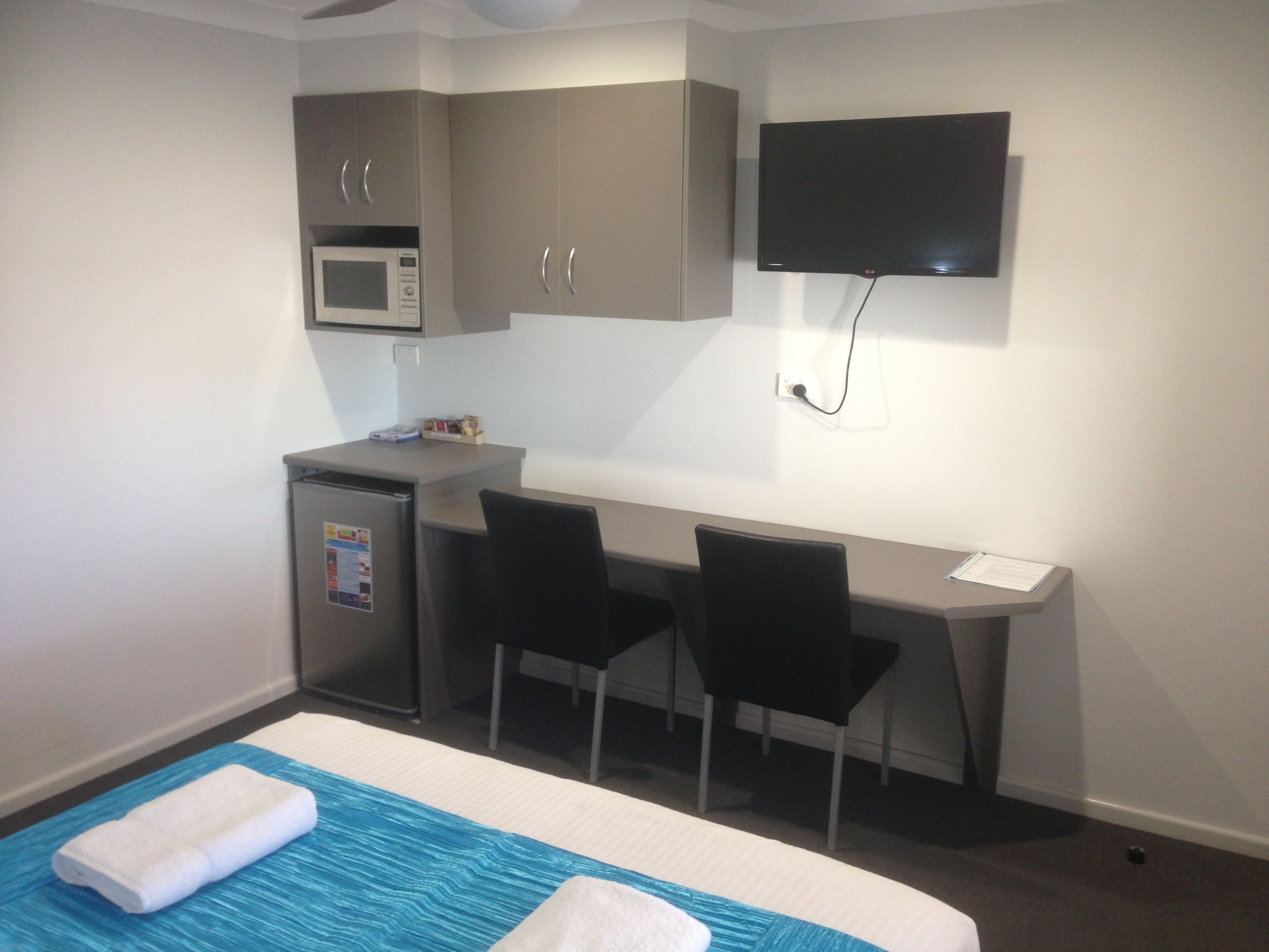 A hotel room with a bed, a desk with two chairs, a microwave, and a flat-screen TV mounted on the wall.