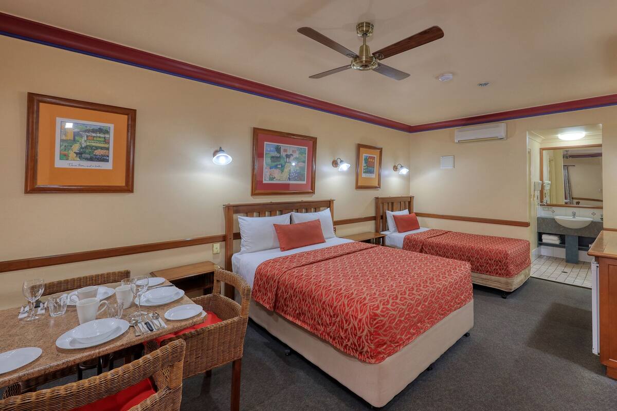 A hotel room with two beds, a dining table with chairs, and a bathroom with a sink and mirror.