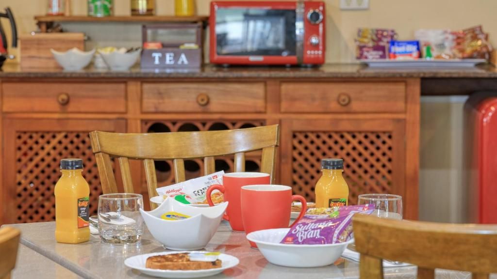 A breakfast table with a bowl of cereal, a red mug, a glass of water, and juice bottles.