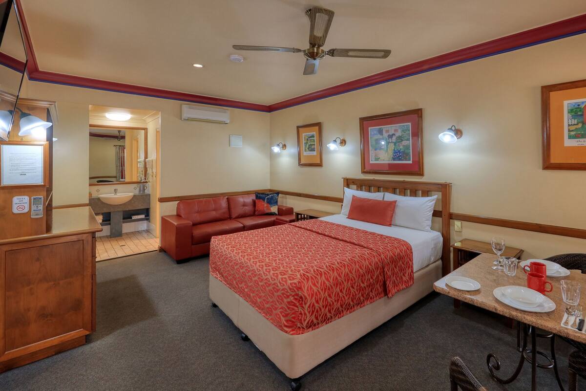 A hotel room with a bed, a sofa, a dining table with plates and glasses, a ceiling fan, and framed artwork on the walls.