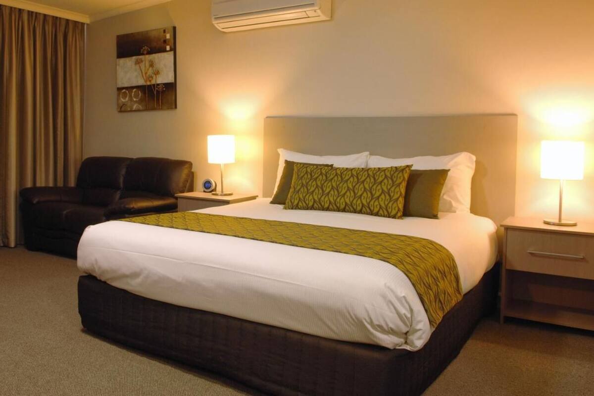 A hotel room with a large bed, two bedside tables, a lamp, a chair, and a wall-mounted air conditioner.