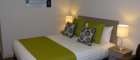 A neatly made bed with a green throw blanket, two pillows with a circular pattern, and a bedside lamp on a nightstand.