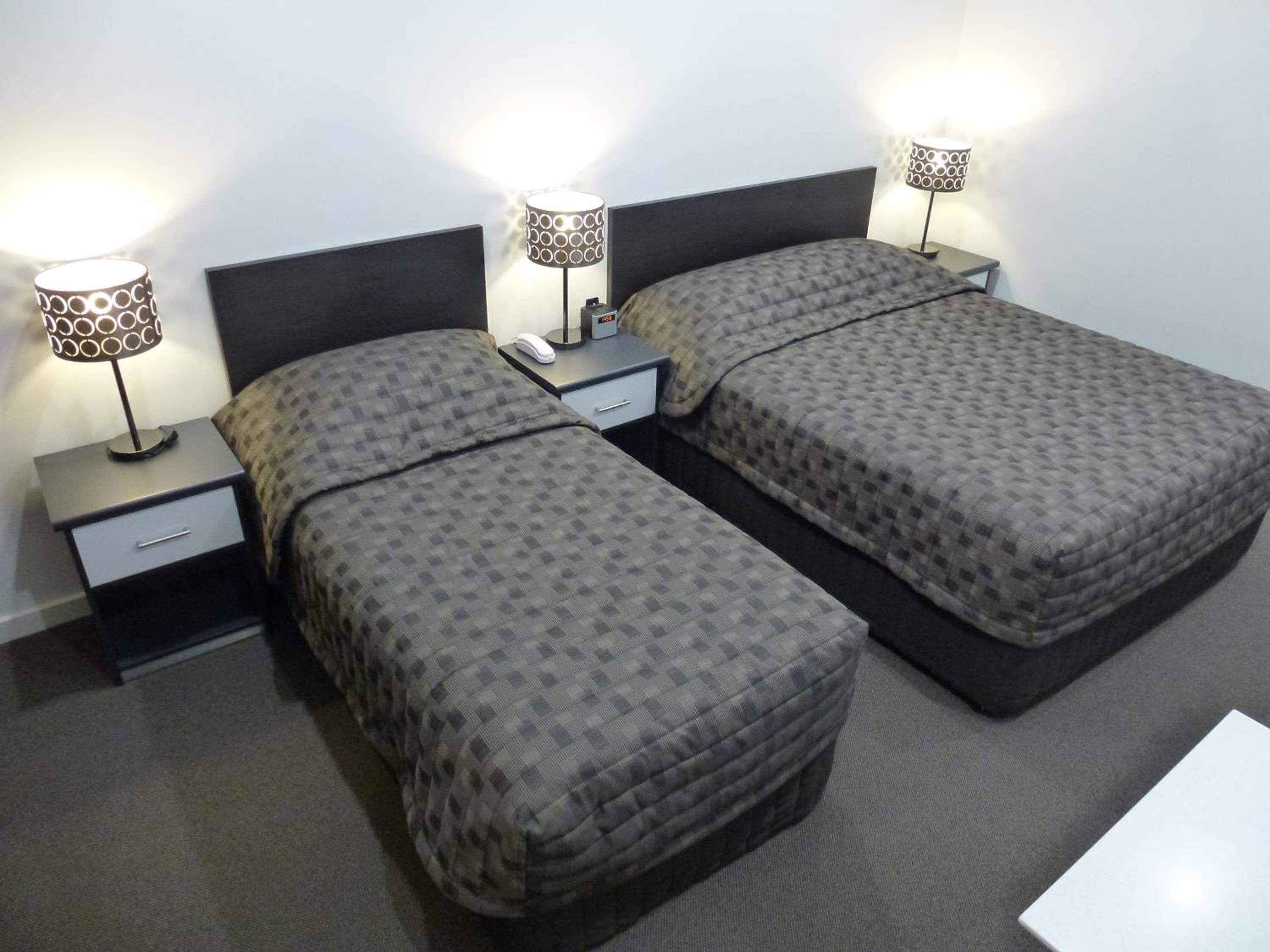 A hotel room with two beds, each with a patterned bedspread, a bedside table with a lamp, and a small drawer.