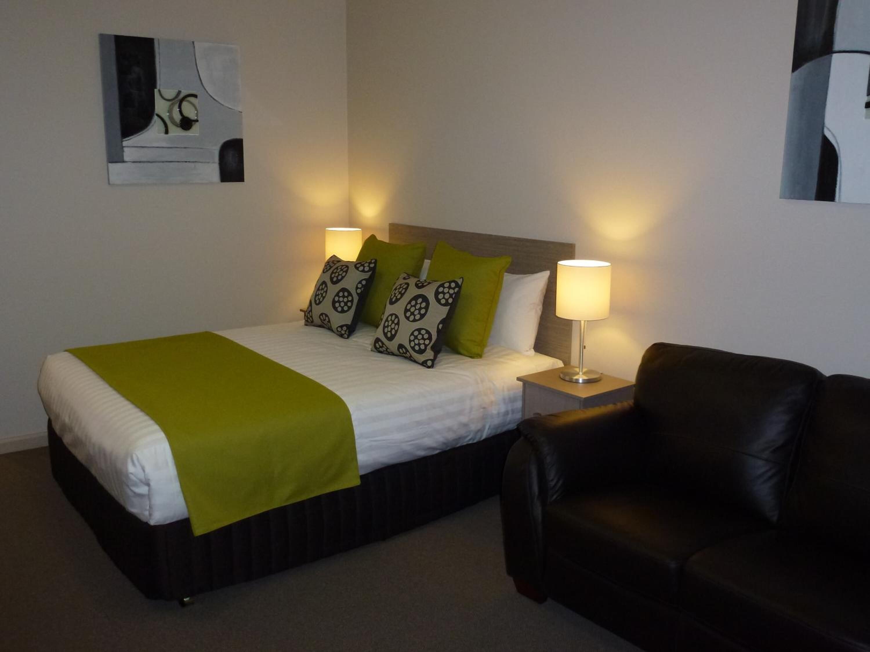 A hotel room with a bed, two bedside lamps, a black leather sofa, and a wall-mounted abstract art piece.