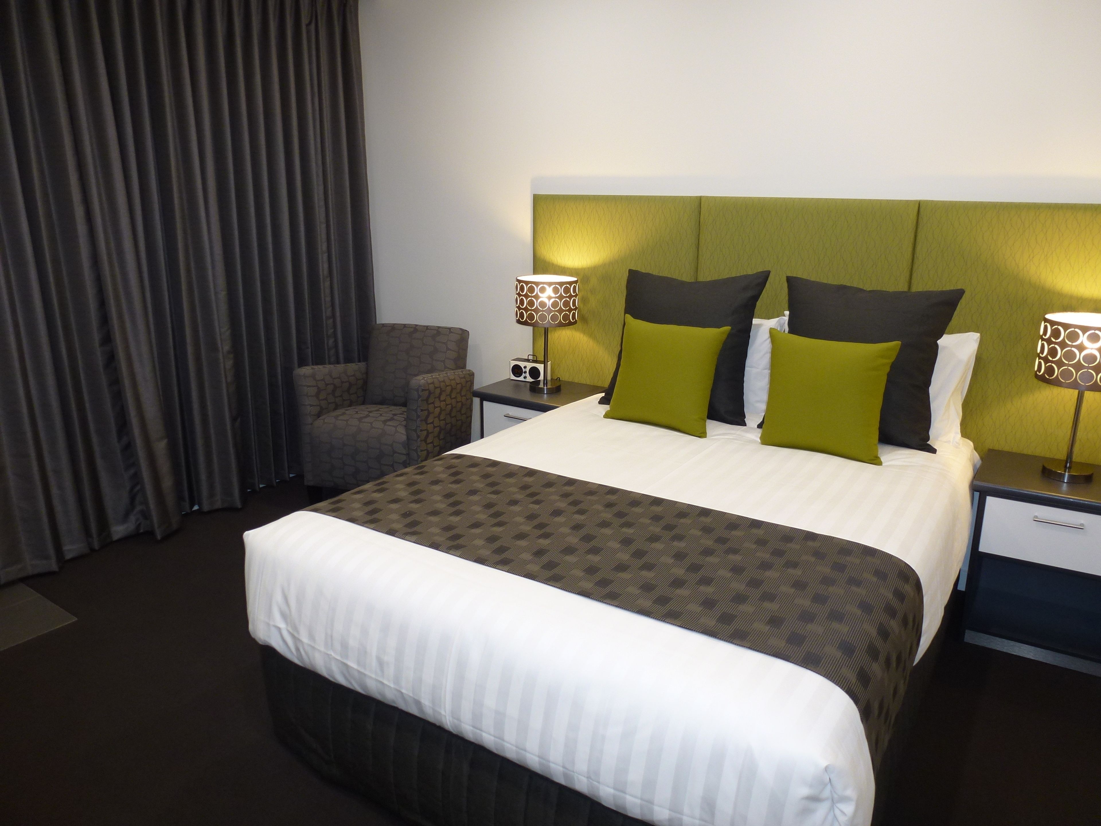 Una habitación de hotel con una cama grande, dos mesitas de noche con lámparas, una silla y una cortina.