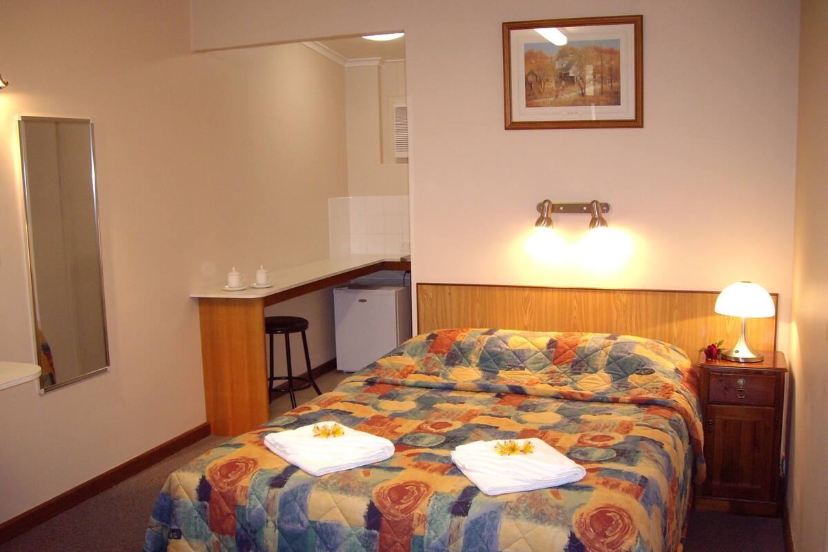 A hotel room with a bed, a desk, a chair, a small refrigerator, a nightstand with a lamp, and a painting on the wall.