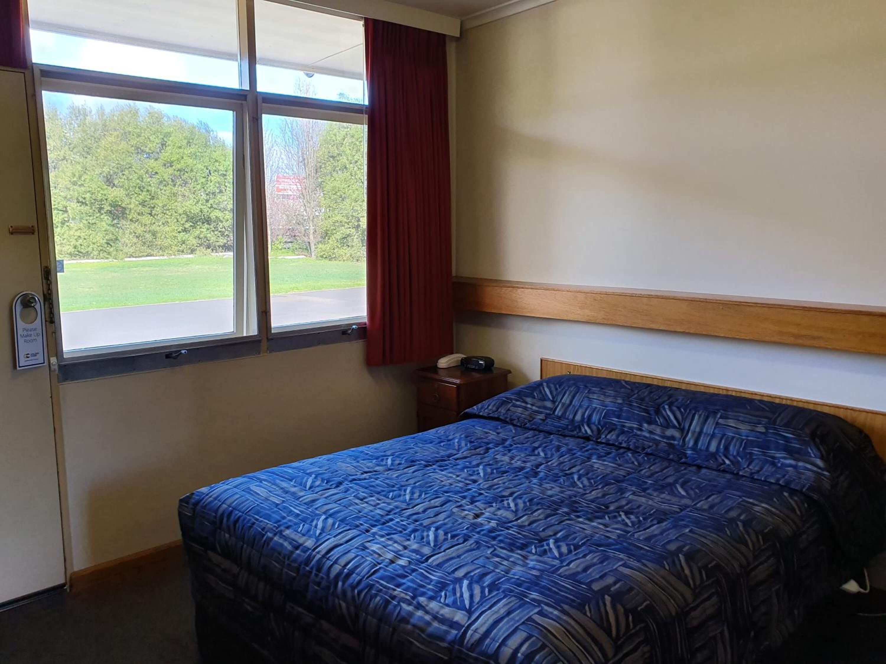 A hotel room with a bed, a nightstand, a telephone, and a window with a view of a green landscape.