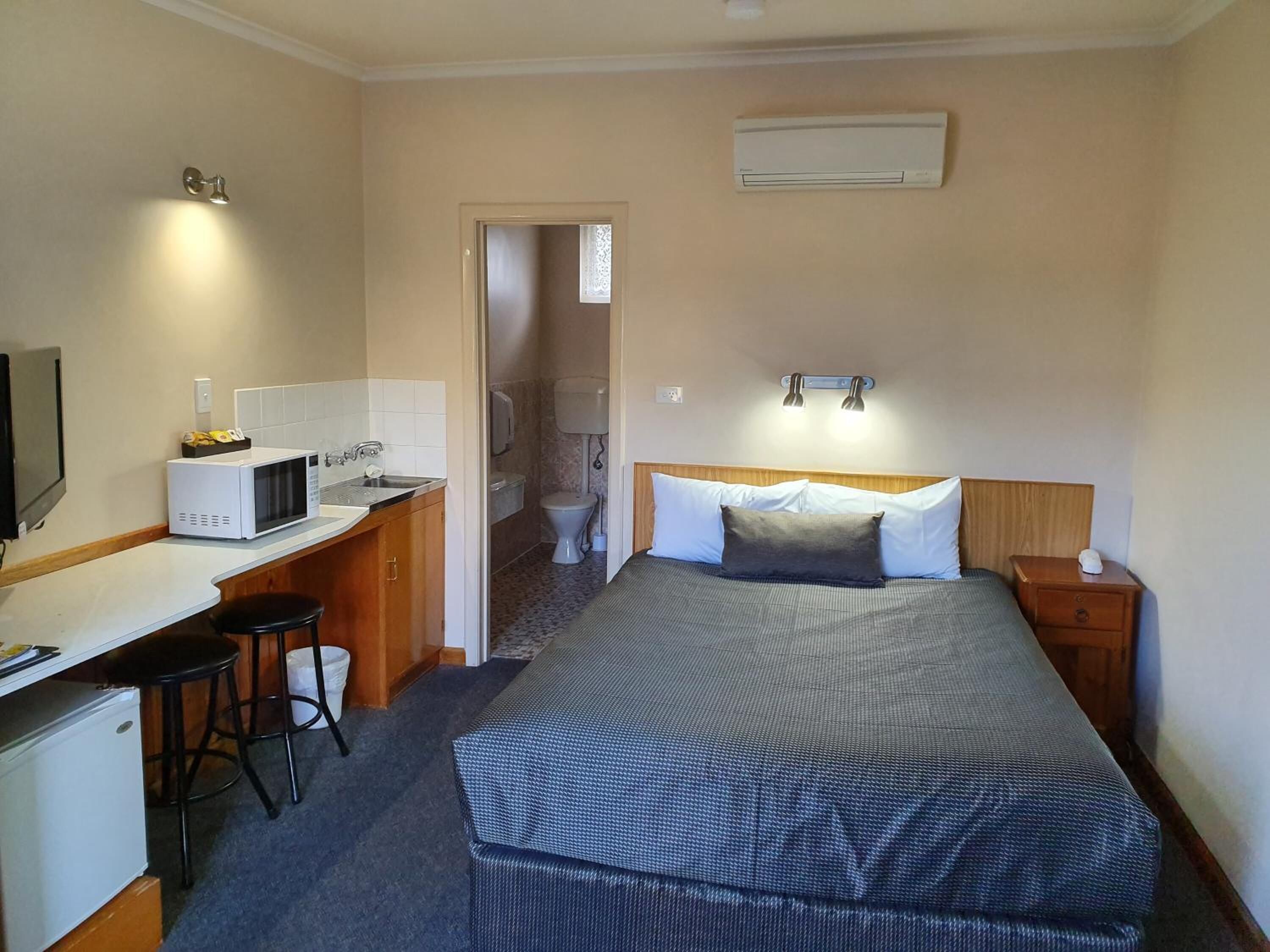 Habitación de hotel pequeña con kitchenette, cama con cabecera, mesita de noche y baño visible a través de una puerta abierta.