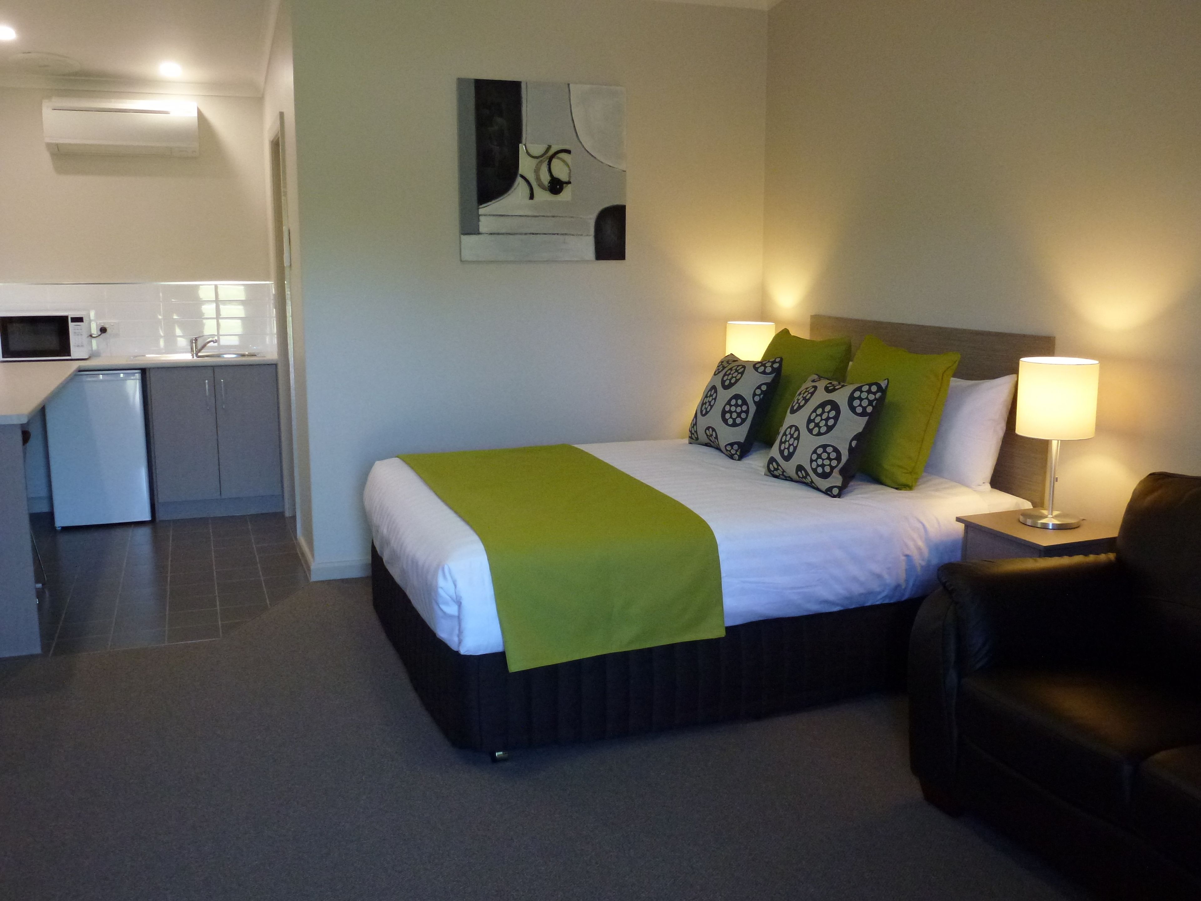 Ein Hotelzimmer mit einem Bett, Kissen in Grün- und Schwarzmuster, einer Nachttischlampe, einem schwarzen Ledersessel und einer Küchenzeile mit Mikrowelle und Spüle.