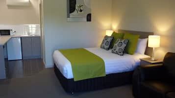 Ein Hotelzimmer mit einem Bett, Kissen in Grün- und Schwarzmuster, einer Nachttischlampe, einem schwarzen Ledersessel und einer Küchenzeile mit Mikrowelle und Spüle.