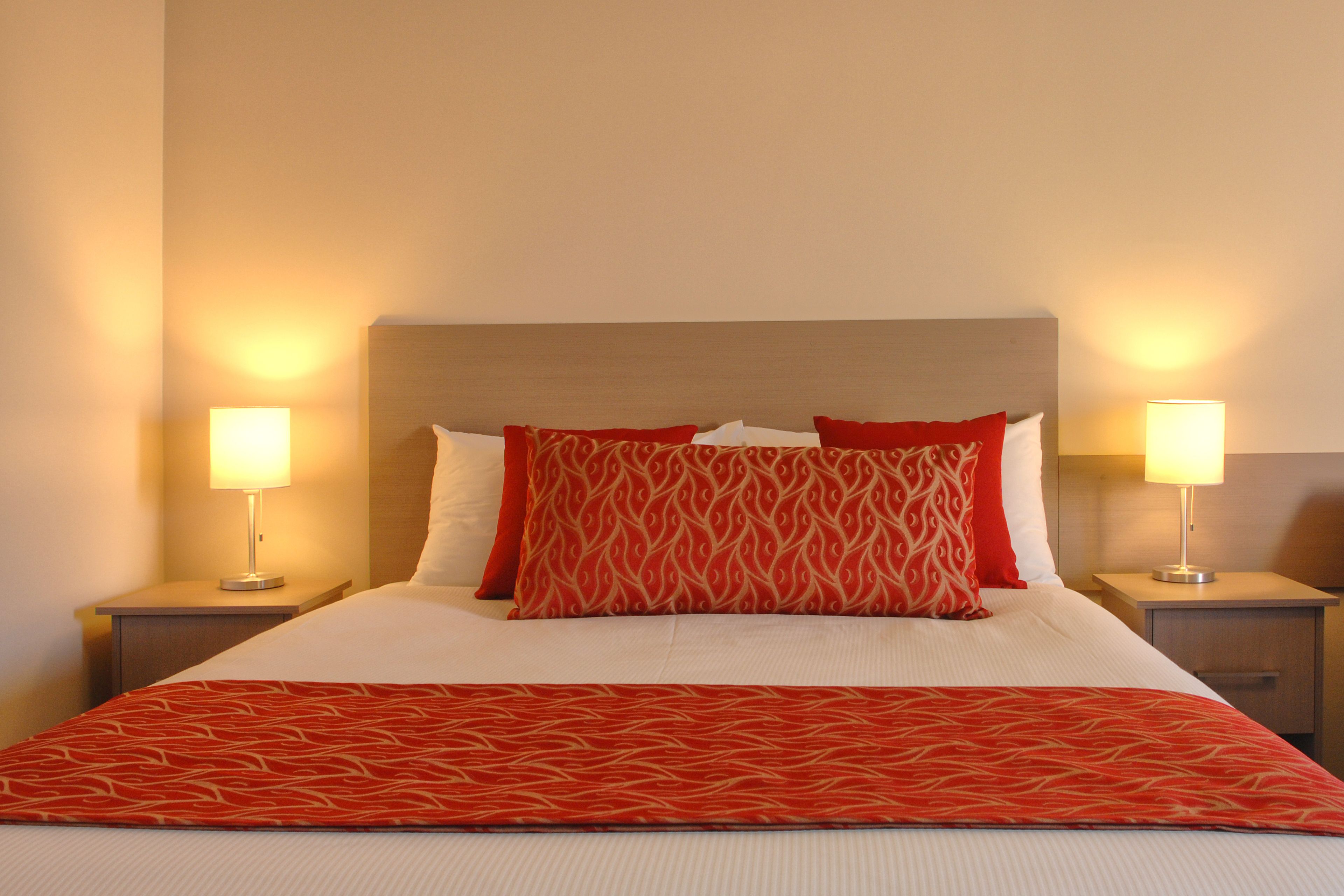 Una cama bien hecha con un edredón rojo estampado, dos lámparas de noche y un cabecero.