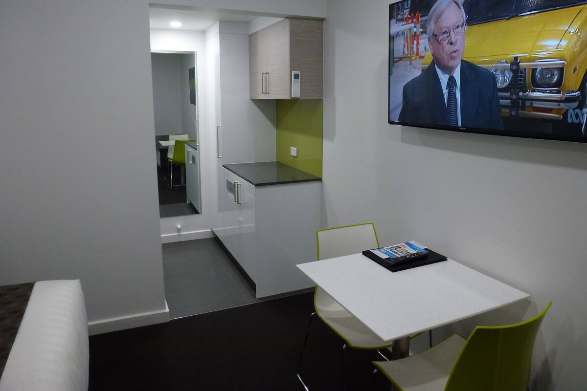 A modern hotel room with a television mounted on the wall displaying a news broadcast, a small table with a magazine, and a green chair.