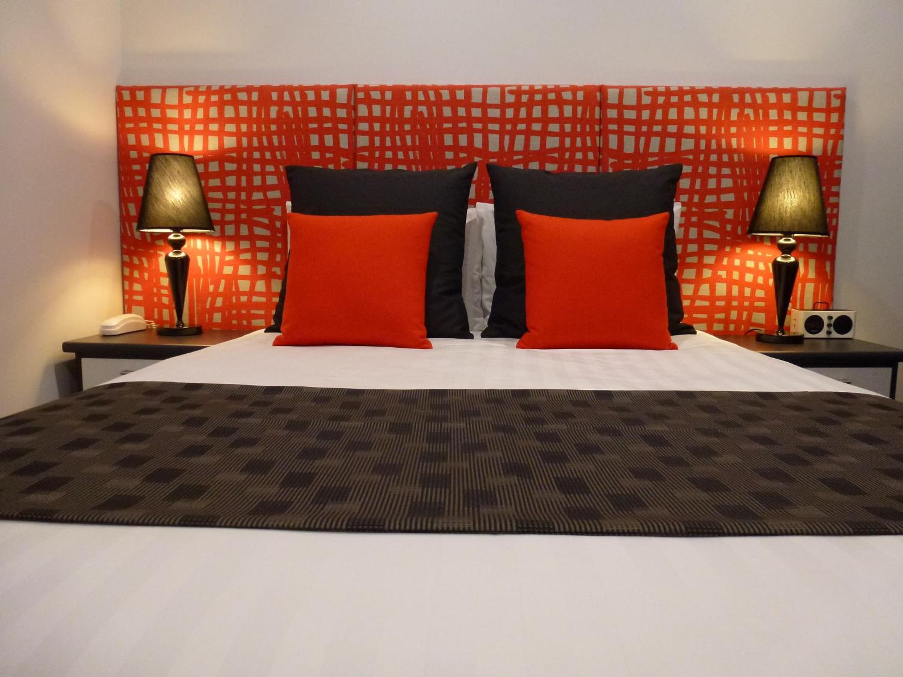 Een hotelkamer met een groot bed, twee rode en zwarte kussens, een beddengoed met een patroon, en twee nachtkastjes met lampen en een radio.