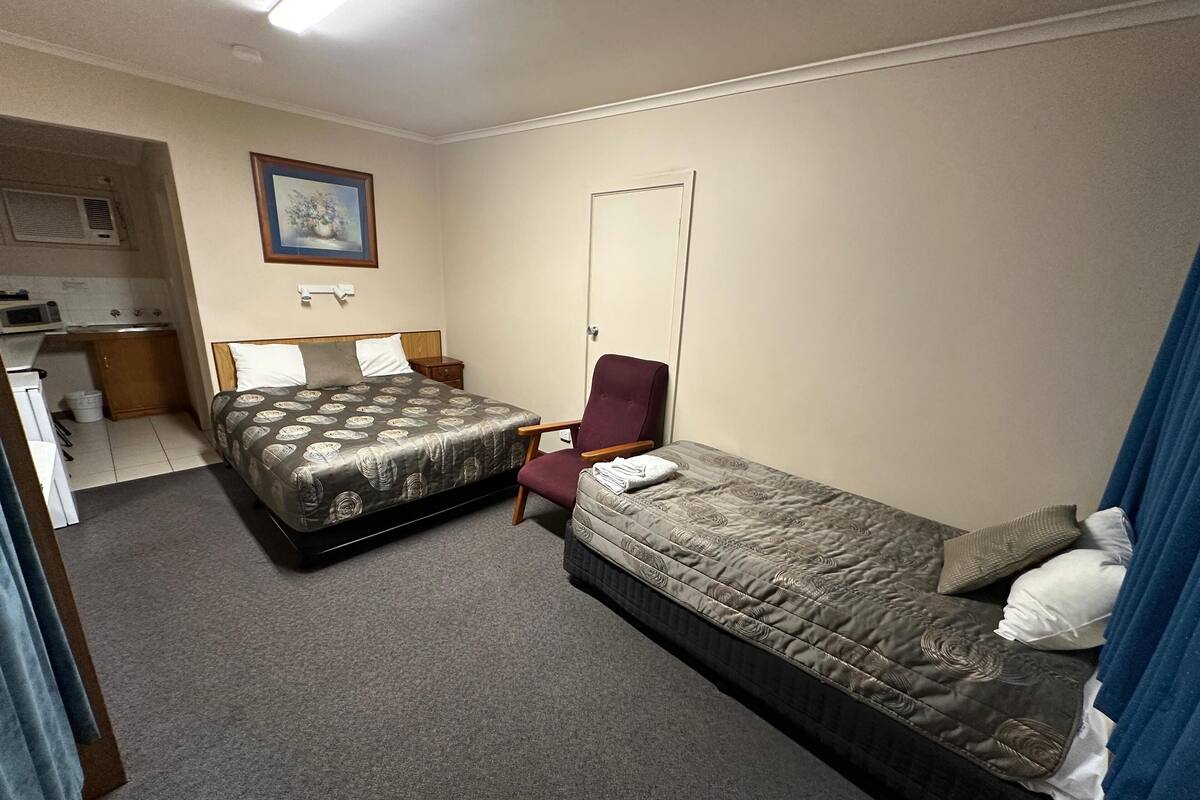 A compact hotel room with a double bed, a single bed, a chair, a desk, and a kitchenette.