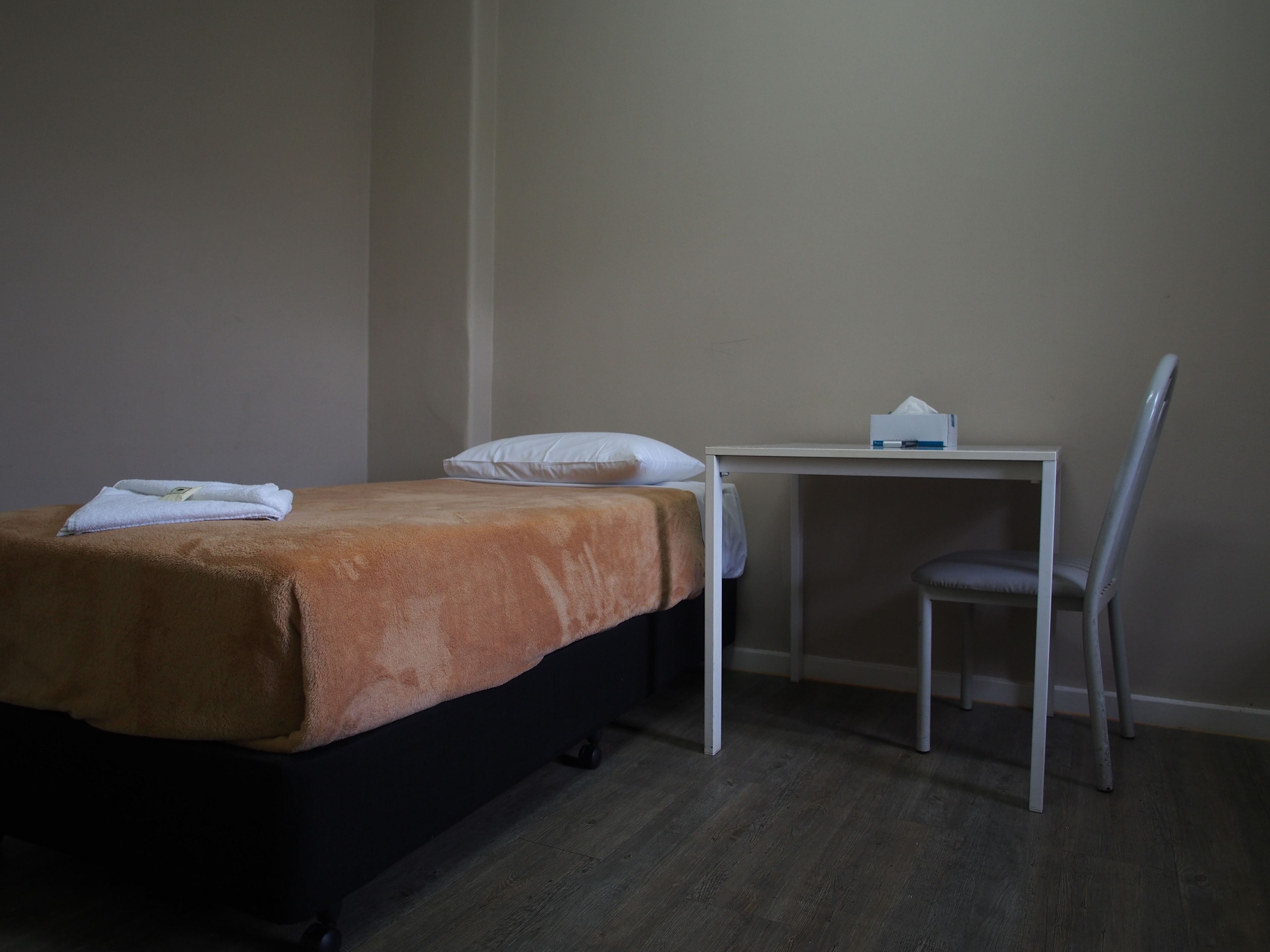 Una cama individual con una manta marrón, un almohadón blanco y una mesita con una caja de pañuelos.