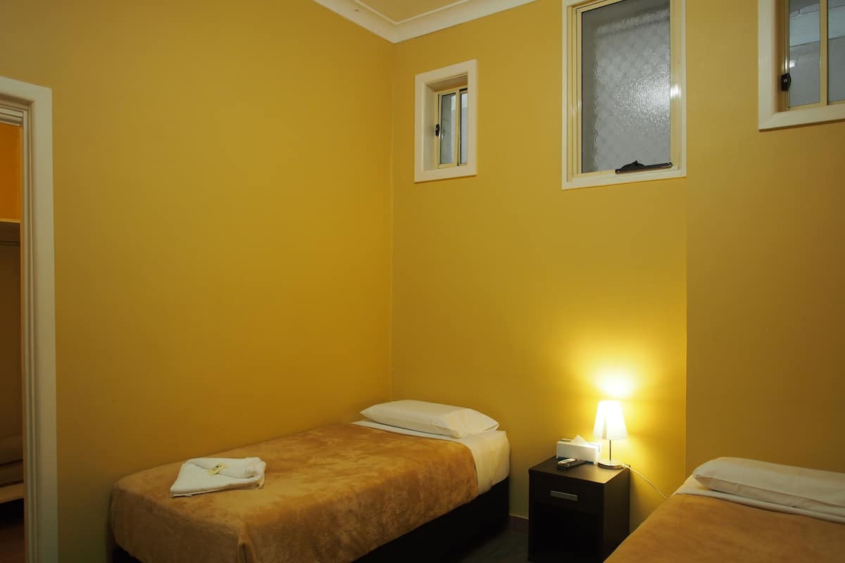 A small, yellow-walled room with two single beds, a nightstand with a lamp, and a window.