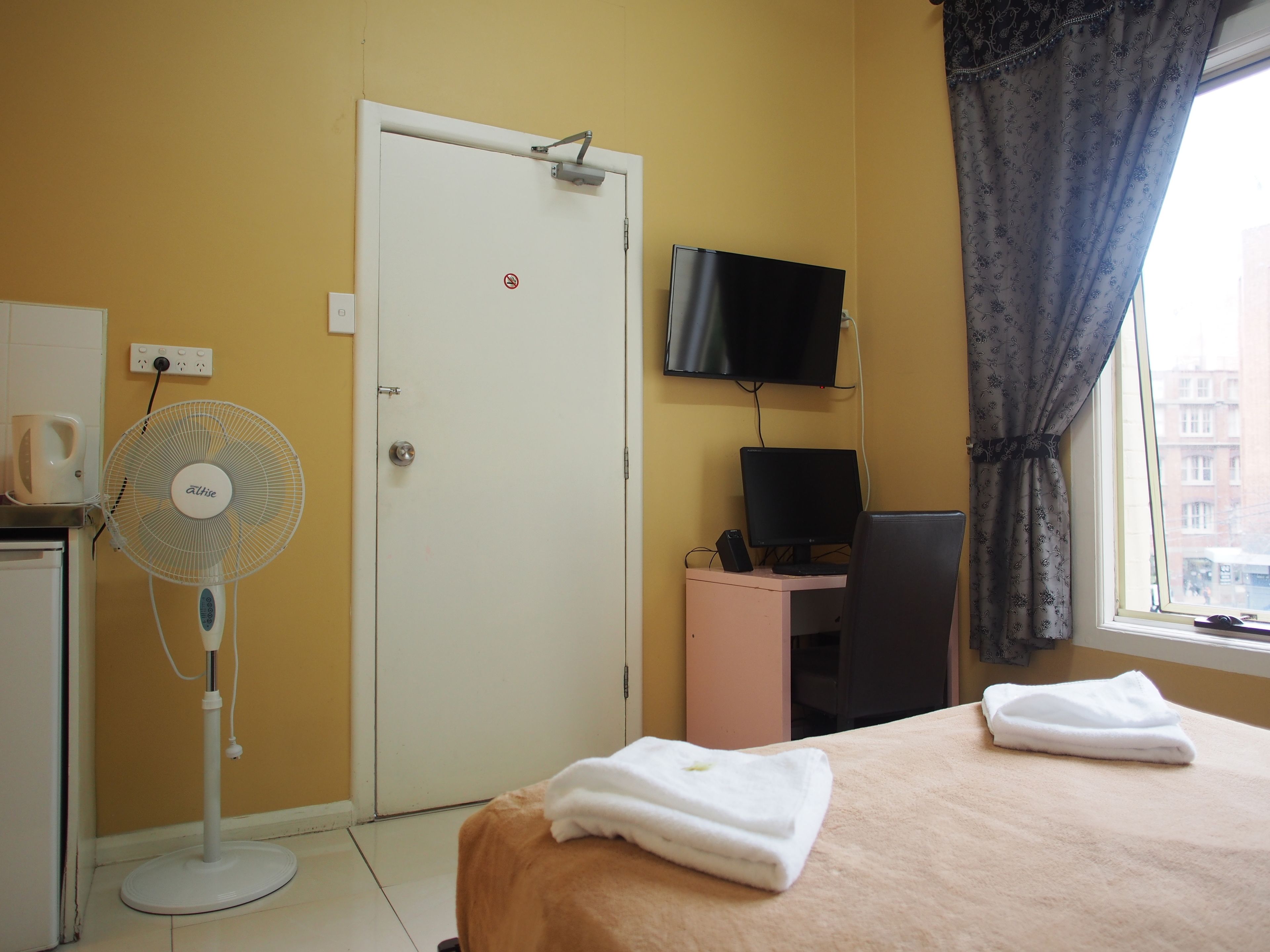 A hotel room with a bed, a desk with a computer, a TV, a fan, and a window with curtains.