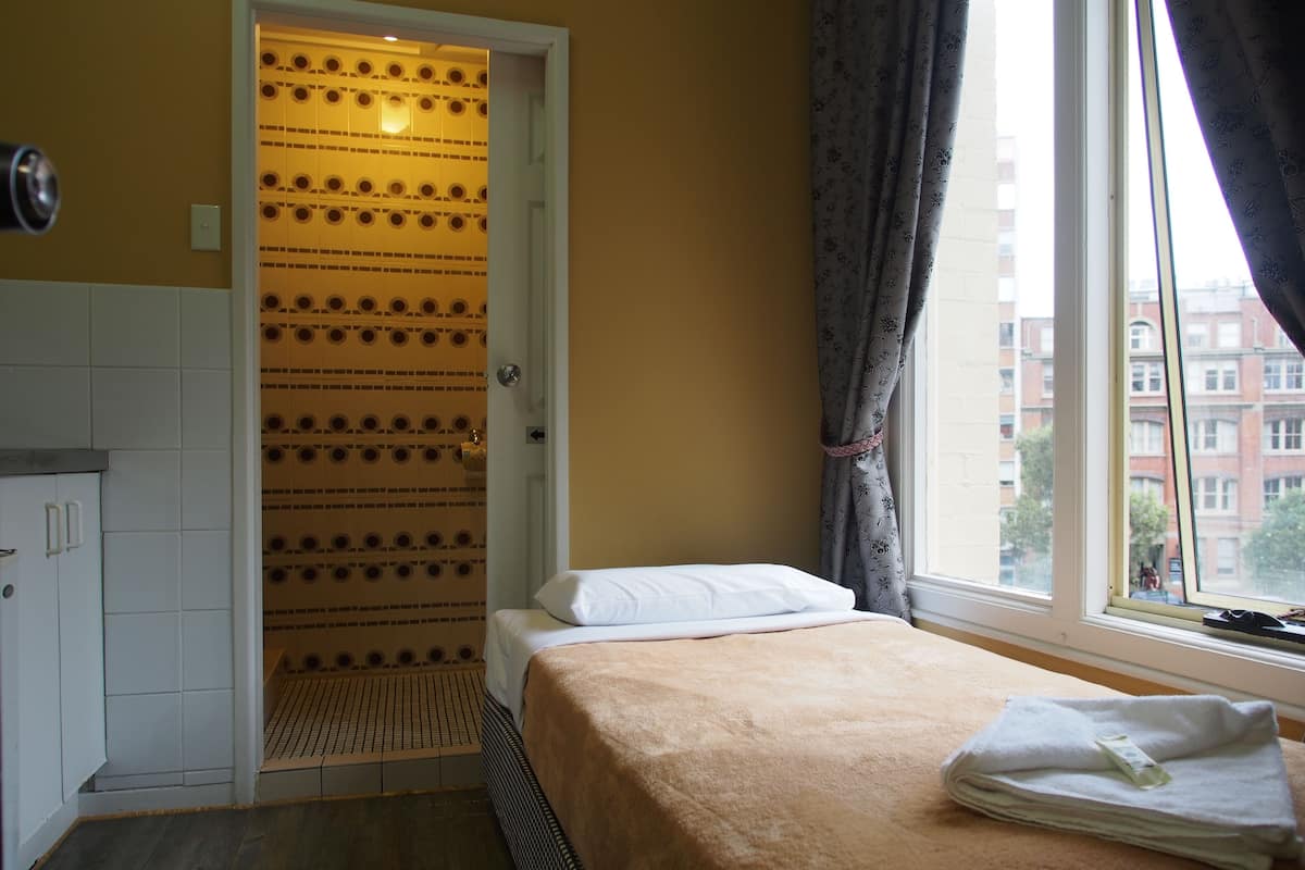 A hotel room with a bed, a window with curtains, a door with a patterned design, and a view of buildings outside.