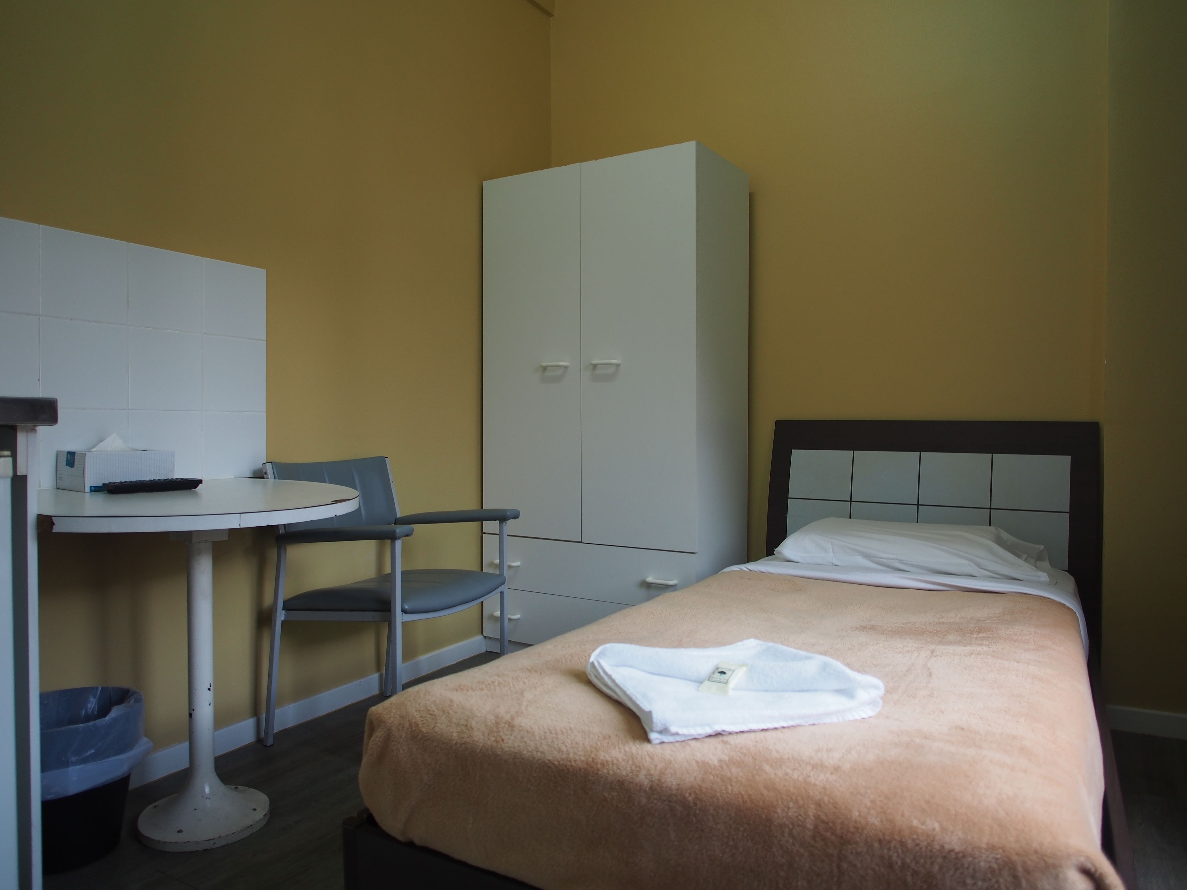 A small, neatly arranged room with a bed, a chair, a table, and a wardrobe.