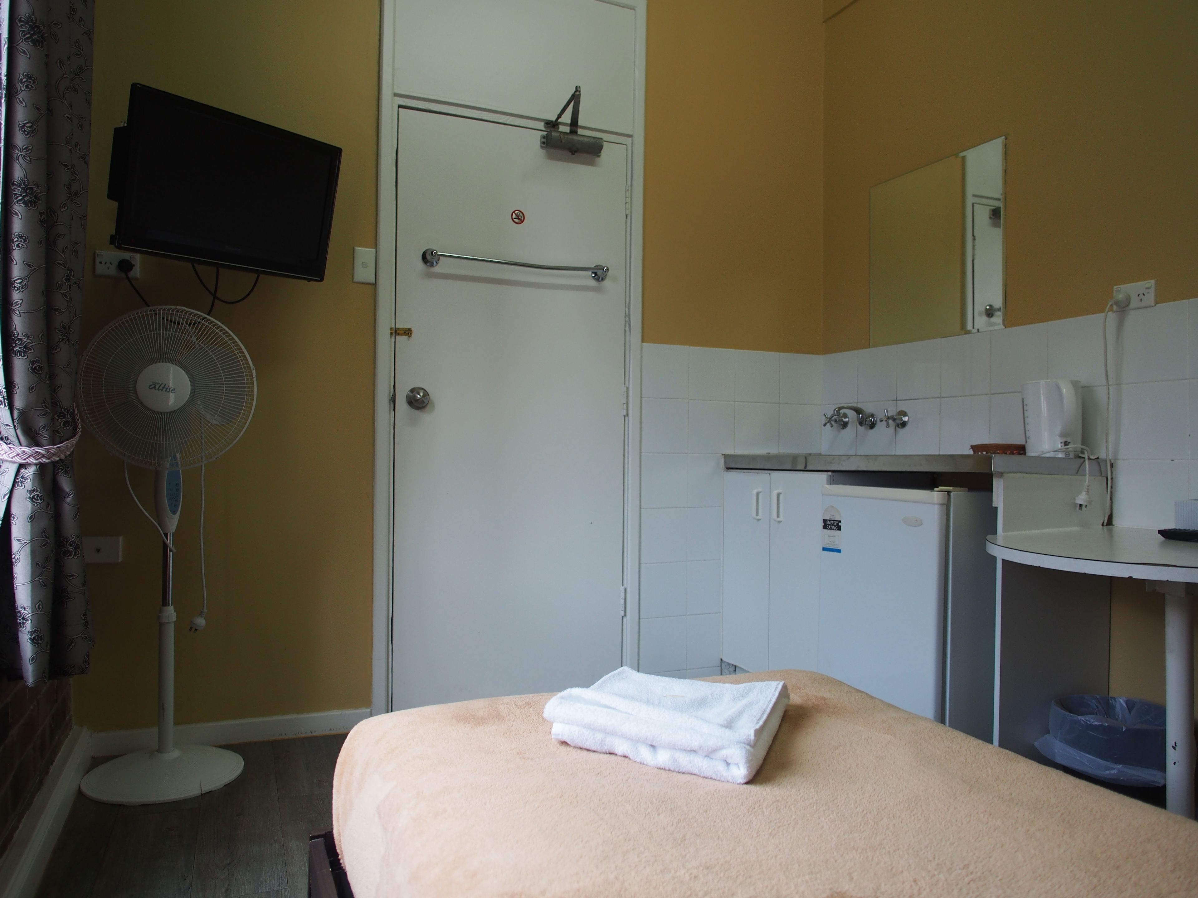 Une chambre d’hôtel comprenant un lit, un téléviseur fixé au mur, un ventilateur, une petite kitchenette et une salle de bain avec douche.